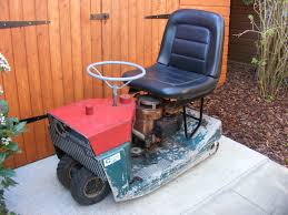 Older red and green ride-on mower with black seat on paving near a wooden shed.